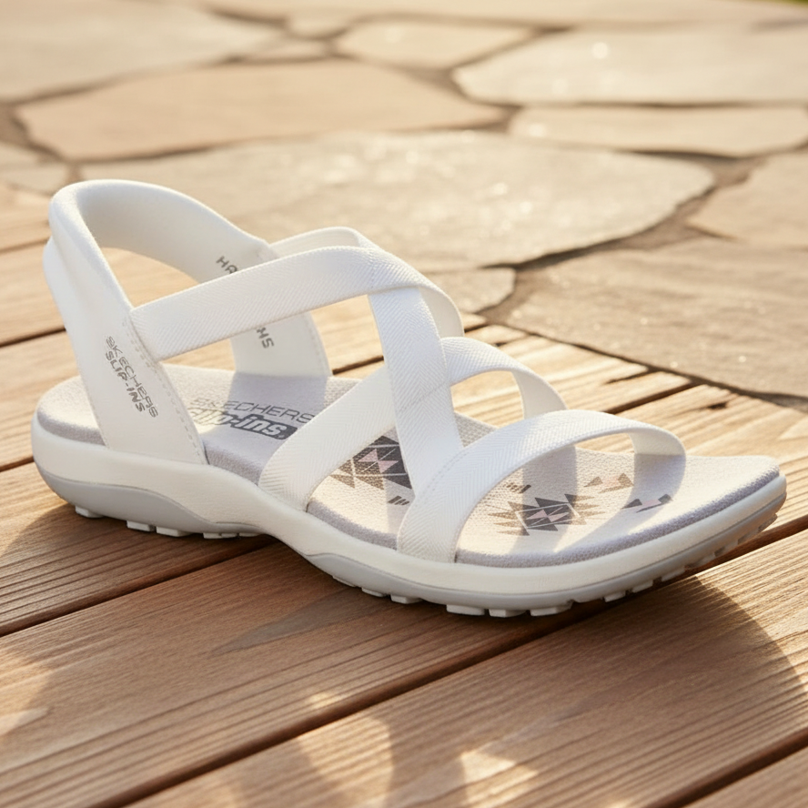 A white women's strappy sandal with a low wedge heel, shown on a wooden deck with stone pavers in the background.