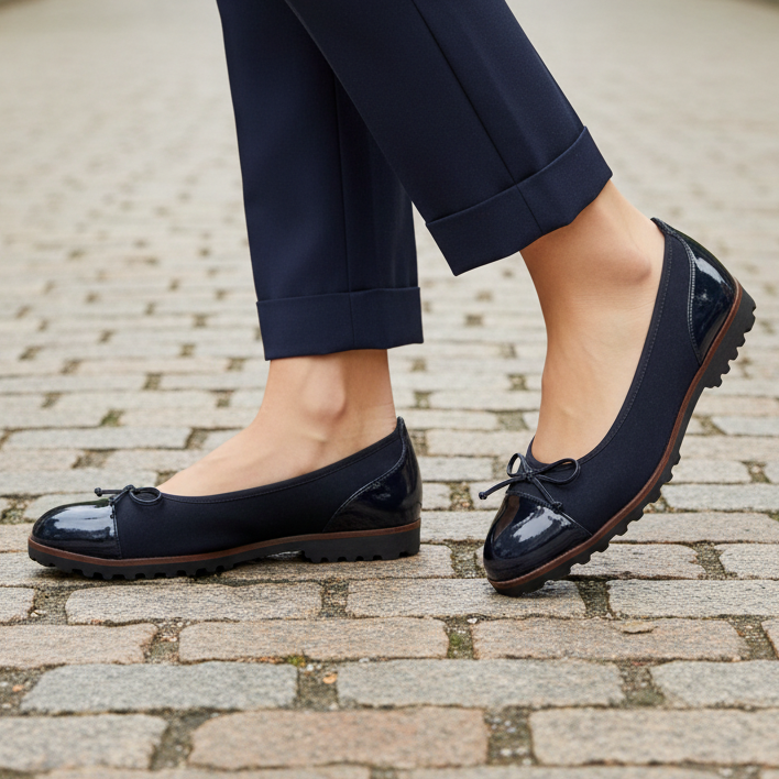 A single women's flat shoe in black patent leather and suede, featuring a small black bow on the vamp and a chunky black lug sole, shown from a side profile against a white background.