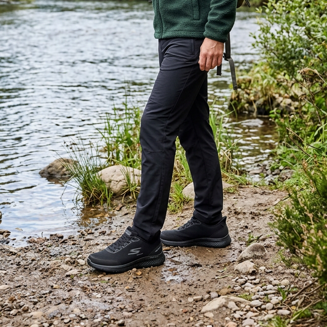 Black Skechers trainers worn on a rocky path beside a stream