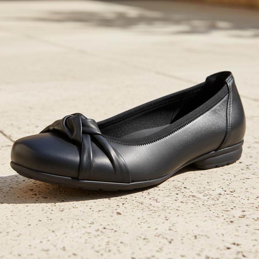 A single black leather ballet flat shoe with a decorative bow on the toe, resting on a light beige stone pavement.