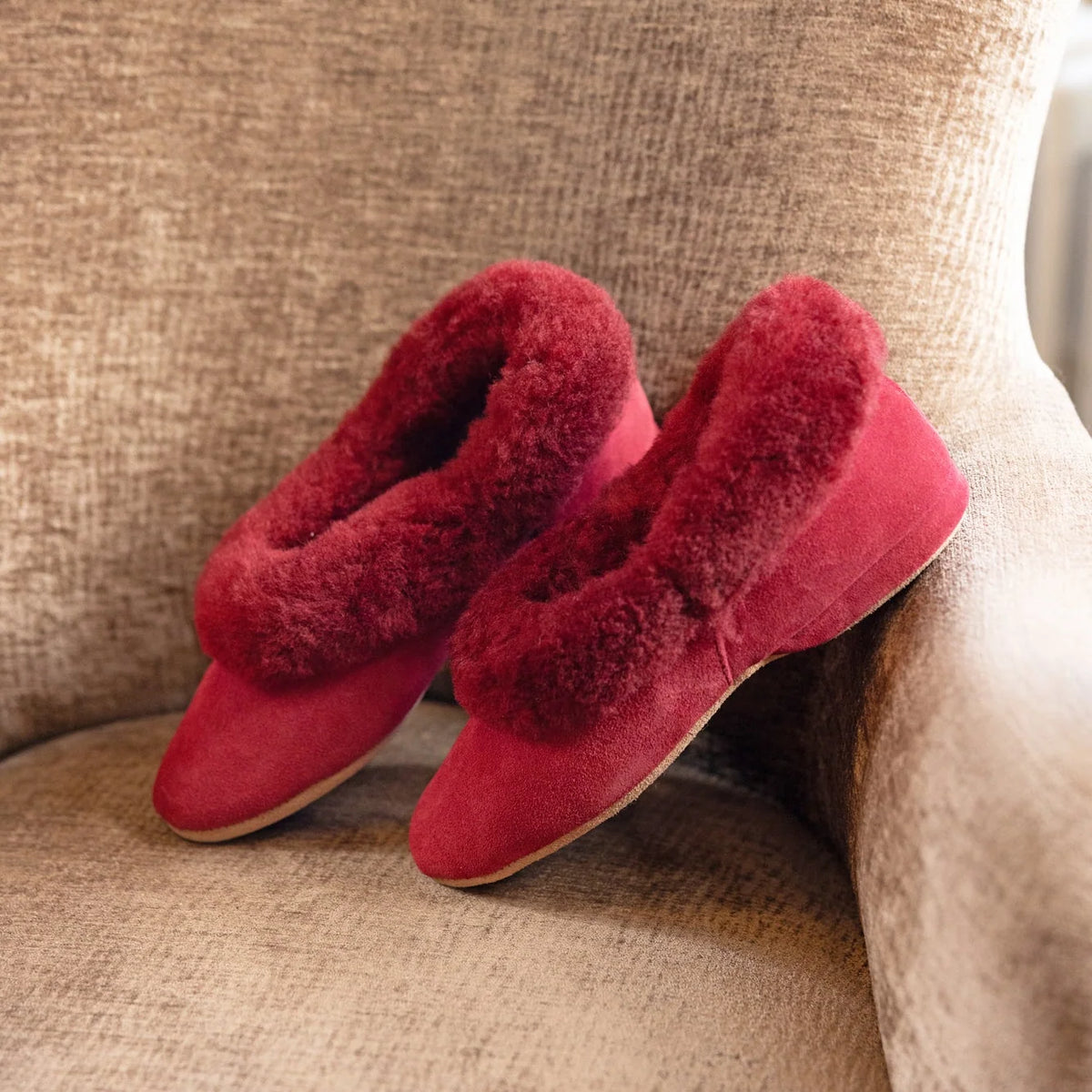 Pair of women’s cherry red sheepskin slippers on a beige couch cushion