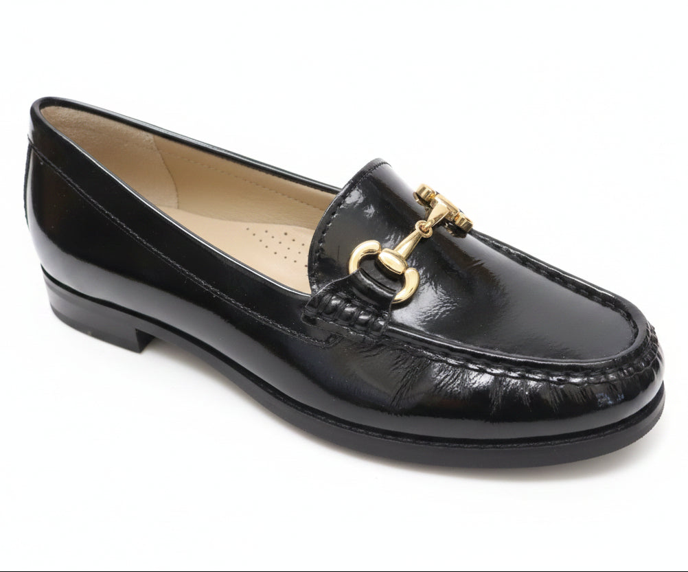 A black patent leather loafer with a gold horsebit detail, resting on a light-colored wooden table.