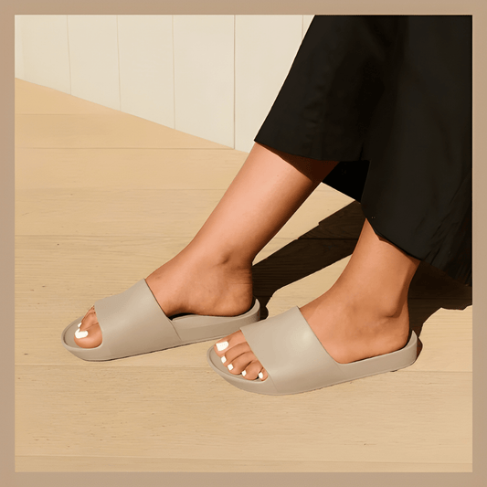 A person wearing taupe open-toe slide sandals with black pants, sitting on a light wood floor against a cream paneled wall.